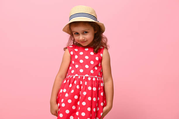 hübsches mädchen in einem rosa sommerkleid mit weißen polka-punkten und strohhut, das schüchtern in die kamera schaut und vor einem rosa wandhintergrund posiert. porträt eines glücklich lächelnden entzückenden kindes 3-4 jahre alt. - 3 4 years cute beauty beautiful stock-fotos und bilder