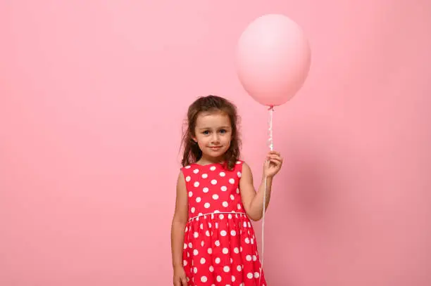 Portrait of beautiful pretty gorgeous adorable 4 years birthday girl, child in dress with polka-dots pattern, holding a pink balloon, isolated on pink background with copy space for advertising.