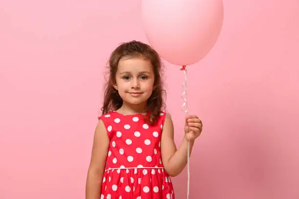 Birthday girl dressed in dress with polka-dots pattern holds a pastel pink balloon, smiles, isolated over pink background with copy space. Close-up portrait of beautiful 4 years child for advertising