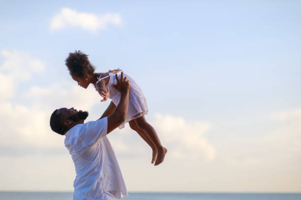 Smiling African American father carrying little daughter on beach during holiday time.