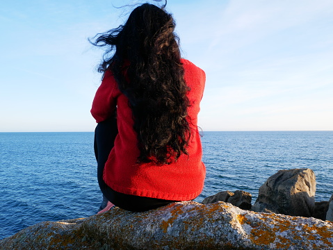Beautiful photo of a woman with long black hair, sitting on a rock, at the edge of the ocean