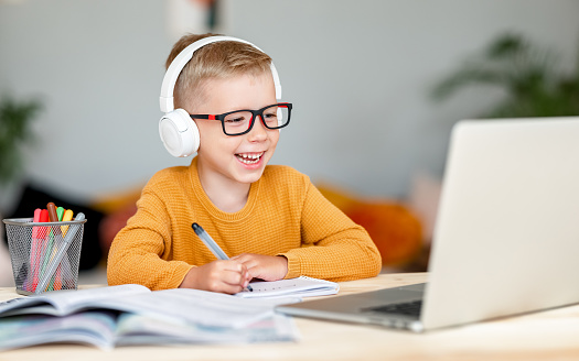 Happy child boy in headphones smiling and looking at screen while making video call to teacher during online lesson at home