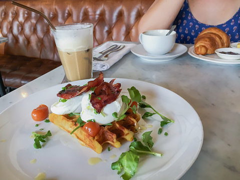Close up food shot of bacon egg waffle above a white plate, with some watercress garnish and cherry tomato. In the background there's one unrecognizable woman sitting on a chair with a croissant in front of her.