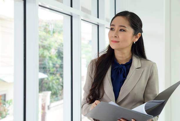 istock Asian female office manager with brown suit holding document folder in her hand. She looked outside while thinking of something. 1334809197