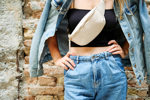 Young beautiful cropped woman standing next to street bricks wall, denim jeans clothes, holding belt satchel purse in shoulder, summer fashion trend, close-up details.
