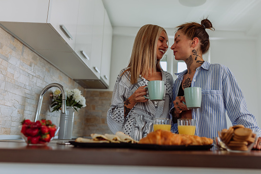 Female same sex couple in pajamas standing at the kitchen counter together having a coffee and a catch up