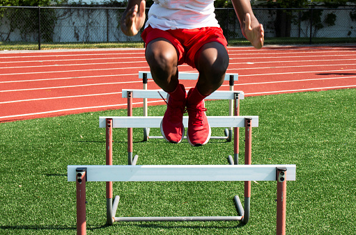 Young male runner training and jumping over hurdles at indoors stadium running track. Sport and competition concept.