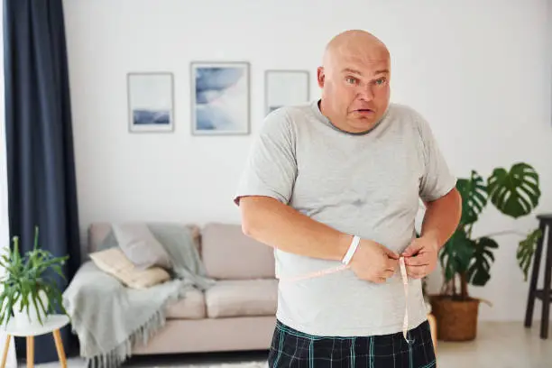 Measuring waist. Funny overweight man in casual clothes is indoors at home.