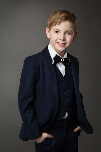 Elegant Little Boy in Classic Black Suit with Bowtie over Gray. Smiling Child Gentleman in Smart Casual Clothing looking at Camera. School Kids Fashion