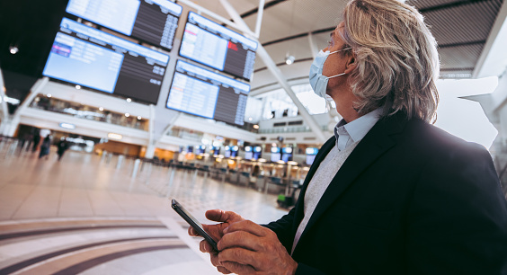 Businessman wearing face mask looking at arrival departure board in airport terminal. Male traveler looking at flight Information screen in the airport during pandemic.