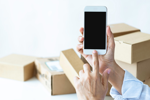 Hand is showing phone screen to camera with ordered gifts in cartoon boxes in background. Representing e-commerce and home delivery.