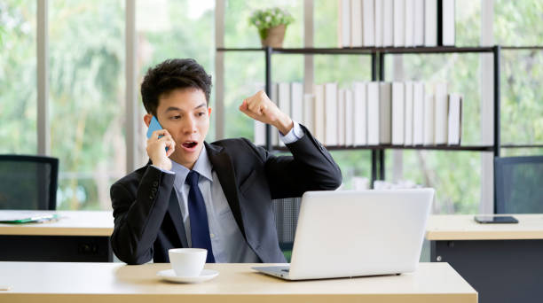 Young man talk to the phone while read email on laptop computer. Asian businessman in suit looking at report excited by good news, personal working with laptop in the office. (yes gesture) joven hablar con el teléfono mientras lee el correo electrónico en la computadora portátil. hombre de negocios asiático en traje mirando informe emocionado por las buenas noticias, personal de trabajo con la computadora portátil en la oficina. (gesto