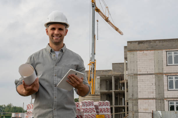 istock Male engineer in a white helmet at a construction site with a tablet computer looks at the camera and smiles 1334837420