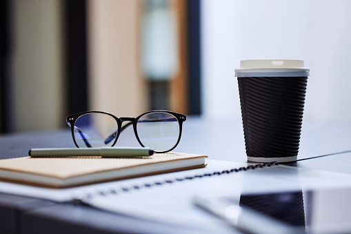 On a sunny day, a notepad for notes lies on the table next to pencils and a laptop. There is a cup of coffee nearby. There is a shadow from the blinds on the table.