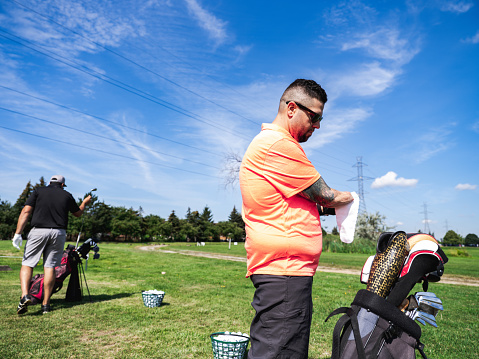Arm amputee and his coach practising golf on the outdoor driving range: Young man with robotic prosthetic lower arm, dressed in sports clothing with another young man dressed the same. Exterior of open field of golf course.