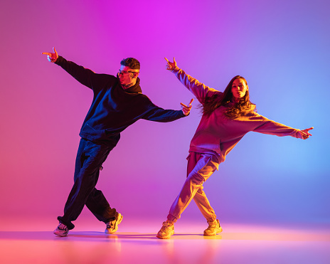 Dos jóvenes, chico y chica con ropa casual bailando danza contemporánea, hip-hop sobre fondo rosa en luz de neón. photo