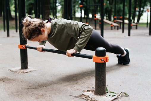 Sporty young fit woman in sportswear exercising on playground doing physical strength exercise, bar push-ups, fitness morning workout outdoors. Healthy active lifestyle concept.