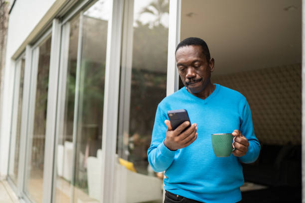 пожилой мужчина смотрит на смартфон дома - men iphone internet social networking стоковые фото и изображения