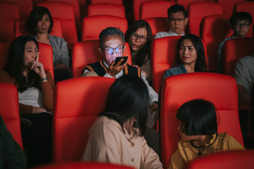 asian chinese senior man talking over the phone loudly  during cinema movie show time in the dark disturbing and ignoring other audience around him. A girl finger on lips asking him to keep quiet