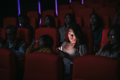 asian chinese woman reading phone message during cinema movie show time in the dark ignoring ansd disturbing other audience around her
