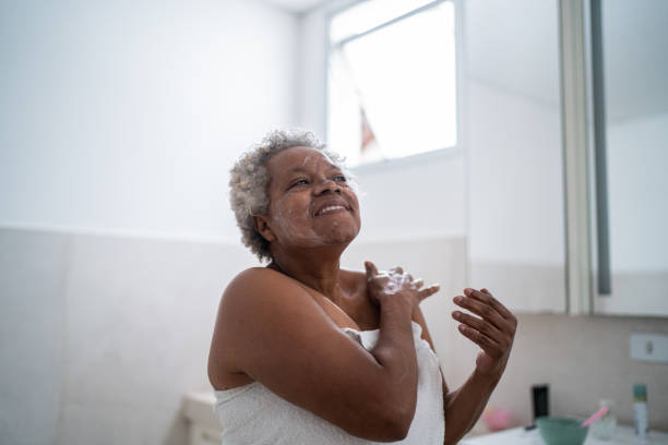 senior woman applying body moisture in the bathroom at home - women body beauty candid imagens e fotografias de stock