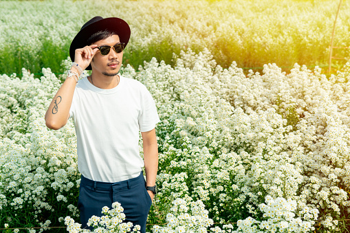 A young Asian man in a white shirt in a black hat stands posing in a white flower garden on a clear day.