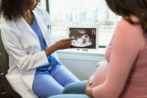 An unrecognizable female doctor points to the ultrasound on the digital tablet as unrecognizable mother looks on.