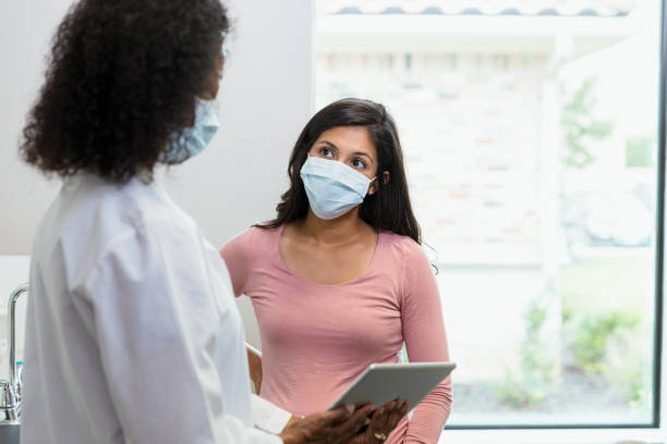Woman with protective mask listens to news given by doctor The mid adult woman wears her protective mask to her appointment because of COVID-19.  She is listening to the information the mature adult female doctor, also wearing a protective mask, is showing her on the digital tablet. protective face mask stock pictures, royalty-free photos & images