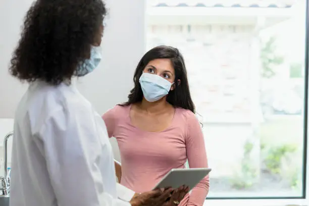 The mid adult woman wears her protective mask to her appointment because of COVID-19.  She is listening to the information the mature adult female doctor, also wearing a protective mask, is showing her on the digital tablet.