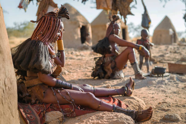 himba-frauen sitzen vor ihren hütten in einem traditionellen dorf in der nähe von kamanjab, namibia, afrika - namibia stock-fotos und bilder