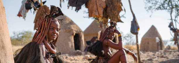 Panoramic View of Himba Women Sitting Outside Their Huts in Traditional Village Near Kamanjab, Namibia, Africa Panoramic shot showing Himba women sitting outside their huts in a traditional Himba village near Kamanjab in northern Namibia, Africa. african tribe stock pictures, royalty-free photos & images