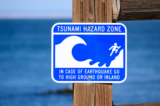 Tsunami Hazard Zone warning sign on ocean coast warn the public about possible danger after an earthquake. Close up. Blurred blue ocean water in background.