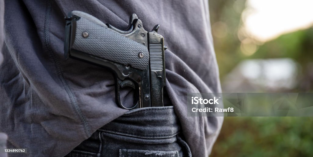 Gloved hand carrying a pistol in his pants, blur nature background, Armed man carrying a pistol in his jeans waistband, blur outdoor nature background. Threat, violence and danger concept Gun Stock Photo