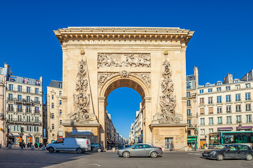 Cars drive past La Porte Saint-Denis in central Paris, France on a sunny day. The arch was commissioned by King Louis XIV.