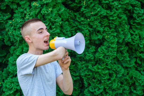 Young caucasian male with amputated arm shouting into a megaphone while standing against green vegetation background