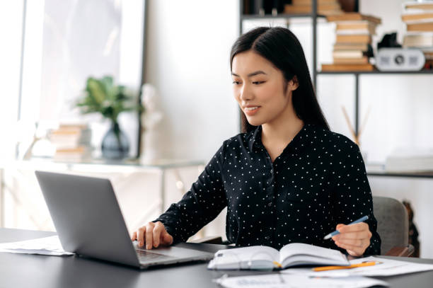 istock Cute smart confident asian girl, freelancer or student sitting at work desk, using laptop for work or study, taking notes during online webinar or distance learning, looking at screen, smiling 1334894911
