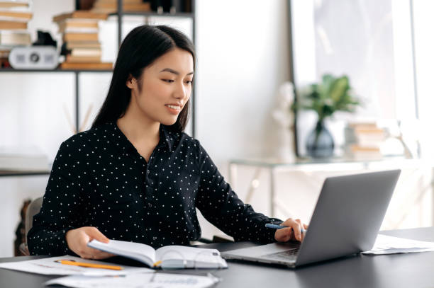 istock Successful beautiful smart young asian woman, freelancer or student sitting at the desk, using laptop for work or online study, taking notes during webinar or online lecture, smiling friendly 1334894912