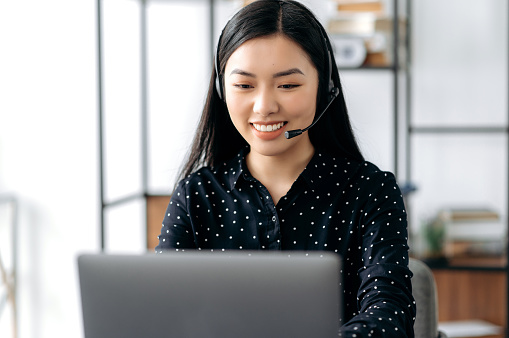 Positive asian young woman, with headset, call center worker or support service operator, sitting at table, using laptop, advising clients, communicate online, smiling friendly