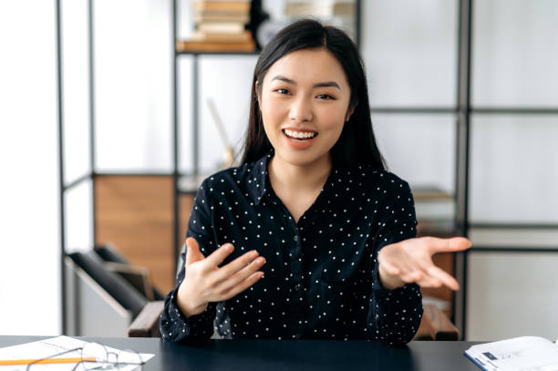 communication vidéo en ligne. sympathique, jolie, intelligente jeune femme asiatique confiante, pigiste ou gestionnaire, s’assoit au bureau, parle avec des collègues ou des amis via une liaison vidéo, gesticulant les mains, souriant - webcam photos et images de collection