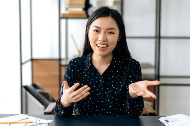Photo of Online video communication. Friendly, pretty, smart confident young asian woman, freelancer or manager, sits at work desk, talking with colleagues or friends via video link, gesturing hands, smiling