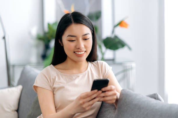 istock Joyful japanese attractive girl using her smartphone while sitting on sofa in stylish clothes, browsing internet and social networks, texting with friends or family, found out good news, smiles 1334894992