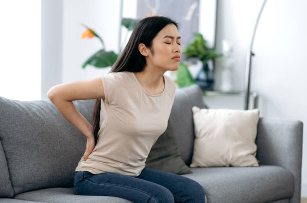 腰痛に苦しむ動揺したアジアの若い女性、リビングルームのソファに座っている不幸な日本のブルネット、背中の痛みのために不快感を感じ、治療と休息を必要とする - arthritis osteoporosis pain backache ストックフォトと画像