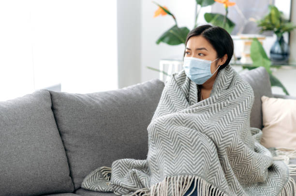 The concept of illness and self-isolation during influenza or covid19. Young sick tired asian woman sits upset on sofa at home, wearing medical protective mask, covered with blanket, looks away sadly stock photo