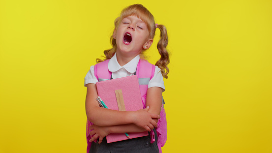 Pretty tired blond teenage student girl dressed in school uniform yawning, sleepy inattentive feeling somnolent lazy bored gaping suffering from lack of sleep. Yellow studio background. Back to school