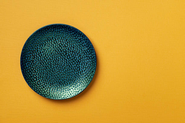 Textured turquoise teal round plate on a yellow textured background. Modern ceramic crockery and tableware for stylish food design. Empty dishes. Copy space.