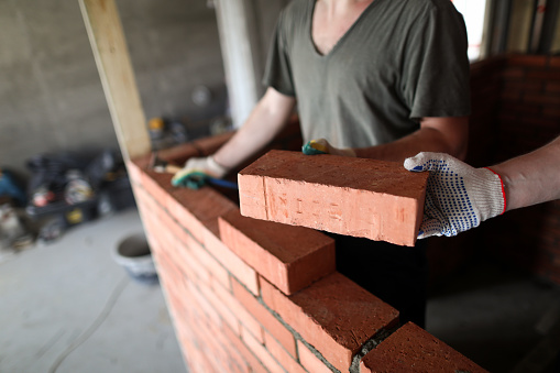 Close-up of construction worker laying red bricks in protective gloves. Handyman building wall with blocks. Man holding hammer. Renovation and professionals concept