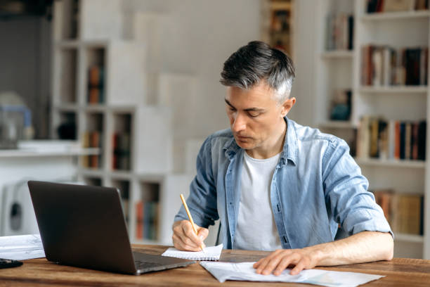 istock Focused smart gray-haired manager, coach or freelancer dressed in casual stylish clothes, working at home with laptop and documents at the table, preparing a business project, taking notes 1334899517