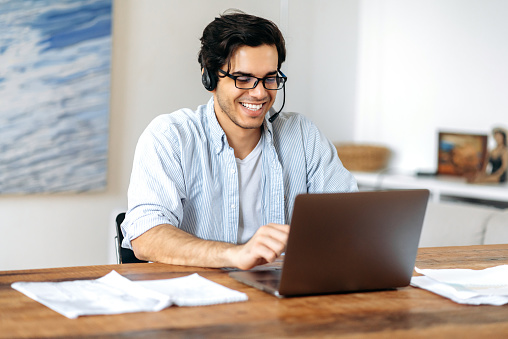 Happy successful confident young hispanic guy in headphones, call center employee or freelancer, working remotely on laptop, communicating via video communication with employees or client, smiling