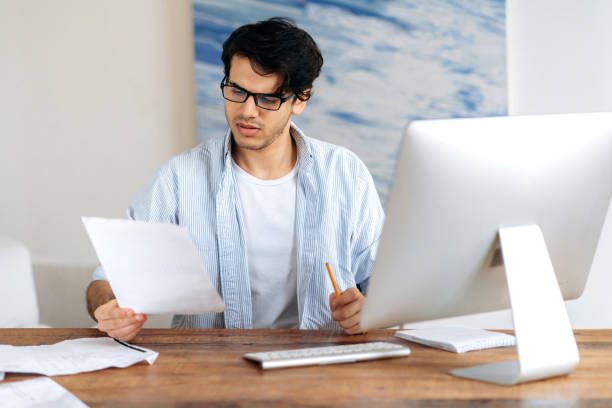 istock Concentrated confident successful handsome hispanic man with glasses, freelancer, student or businessman working or studying rat the desk with computer, looking through business documents 1334899574