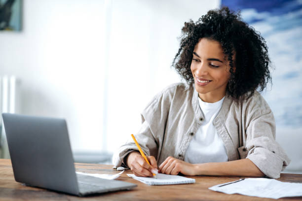 Distance learning, online lesson, work remotely. Focused smart african american female student or freelancer, in stylish wear, studies or works remotely, uses laptop, takes notes during online lecture stock photo
